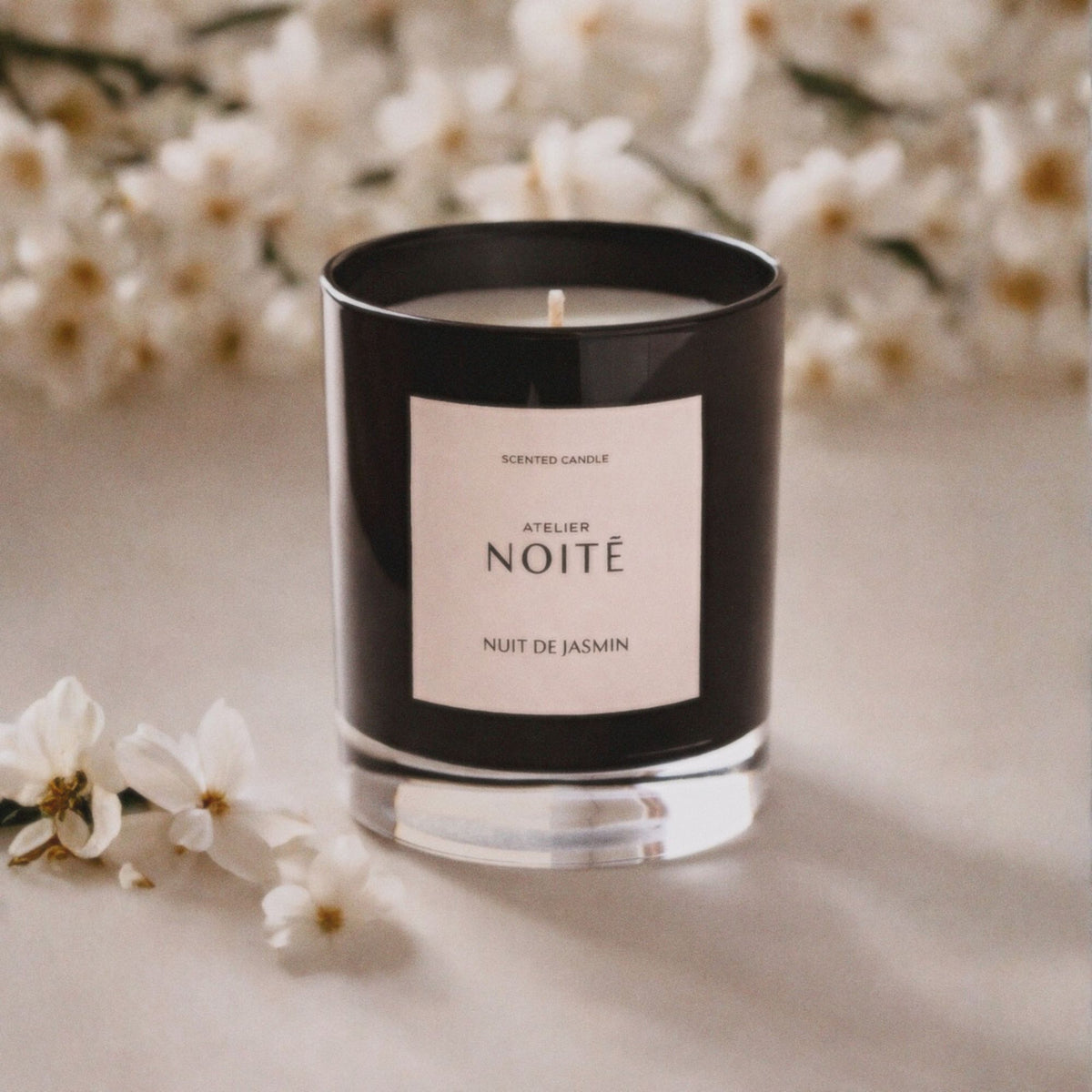 Candle in a black container with 'Atelier Noitē' label on a light surface with white flowers in the background
