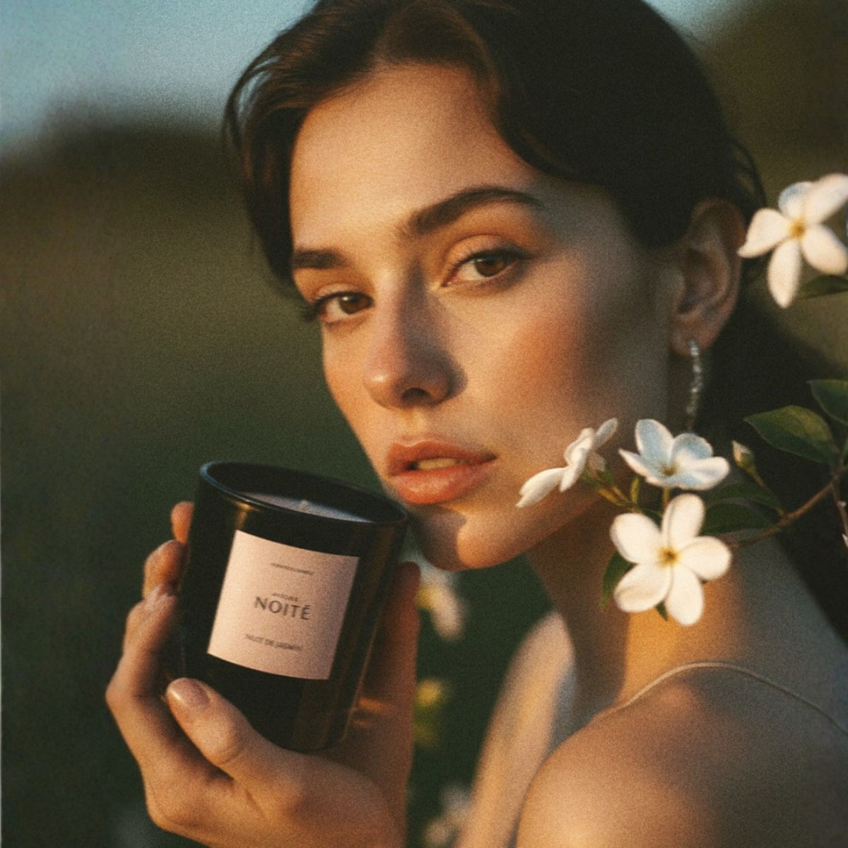 Woman holding Atelier Noitē Nuit de Jasmin Candle with flowers in her hair outdoors.