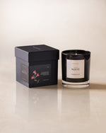 A black candle labeled 'NOITE' with a floral pattern on the packaging, presented next to its box on a light background.
