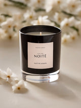 An image of a candle in a black glass jar with a beige label with a beige background and white jasmine flowers