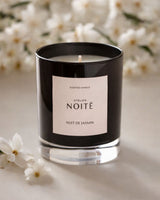 A black glass scented candle jar with a white label reading 'Atelier Noitē NUIT DE JASMIN' on a light surface, surrounded by soft white jasmine flowers.