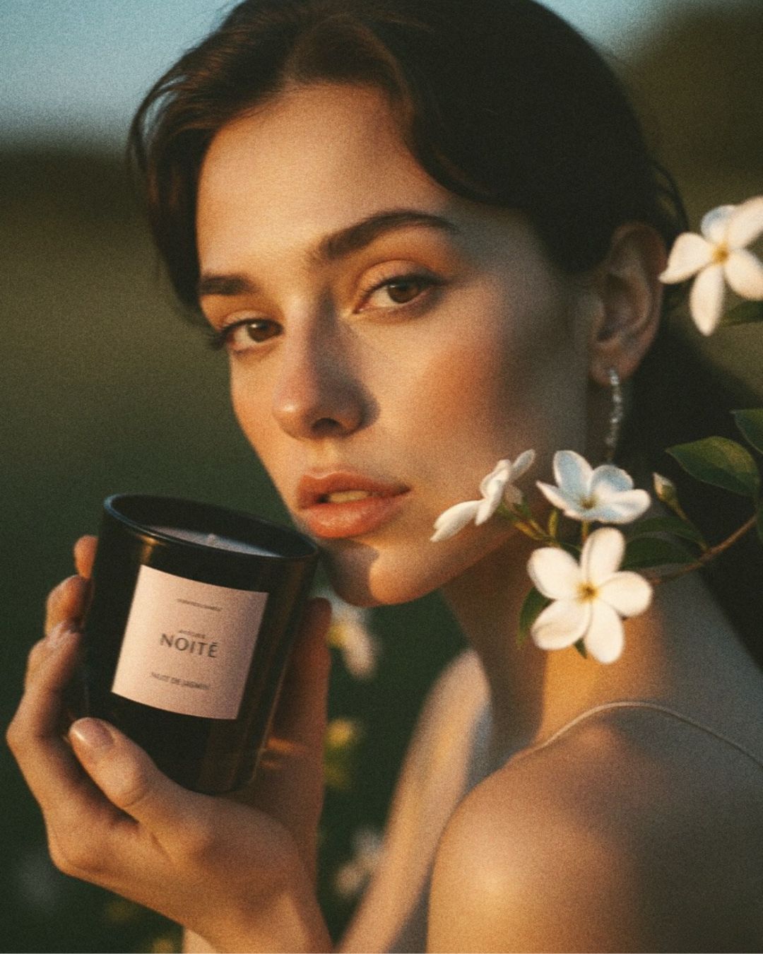 A woman outdoors at golden hour holds a black candle jar labeled 'Atelier Noitē. White flowers and green leaves are near her face, with a softly blurred natural background.