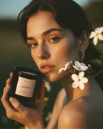 A woman outdoors at golden hour holds a black candle jar labeled 'Atelier Noitē. White flowers and green leaves are near her face, with a softly blurred natural background.
