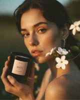 A woman outdoors at golden hour holds a black candle jar labeled 'Atelier Noitē. White flowers and green leaves are near her face, with a softly blurred natural background.