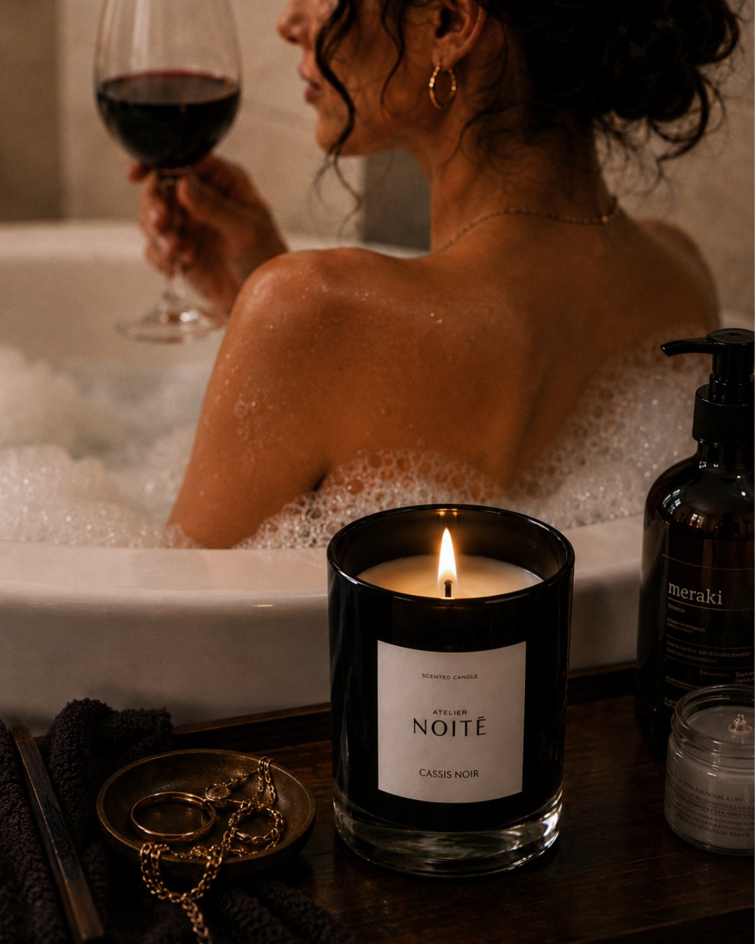 Woman in a bathtub with a candle labeled Atelier Noitē' and a glass of wine.