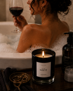Woman in a bathtub with a candle labeled Atelier Noitē' and a glass of wine.