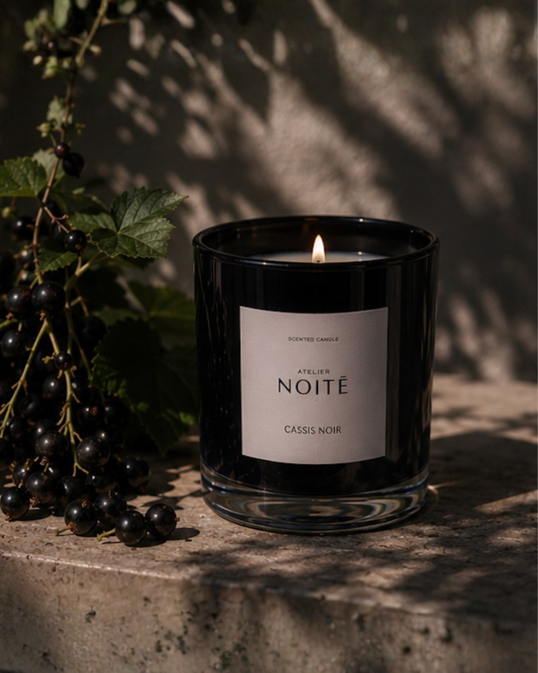 Black candle labeled 'Atelier Noite' with a label on a stone surface next to black currants.