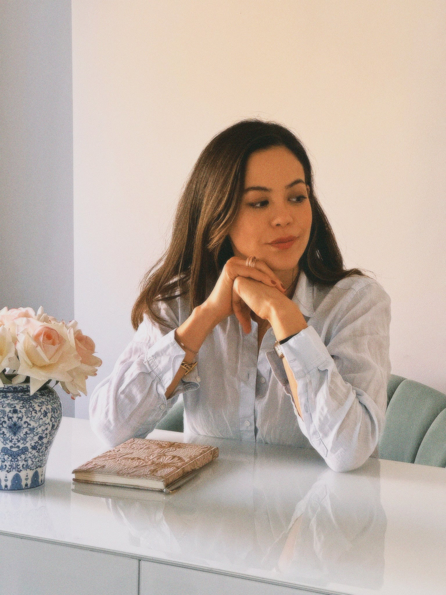 Natalie Schober, founder of Atelier Noite,  wearing a light blue striped button-down shirt. A blue and white patterned vase with pink roses and a textured book are on the table next to her.