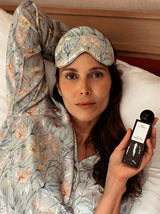 Woman in floral pajamas and eye mask holding a room spray product in bed