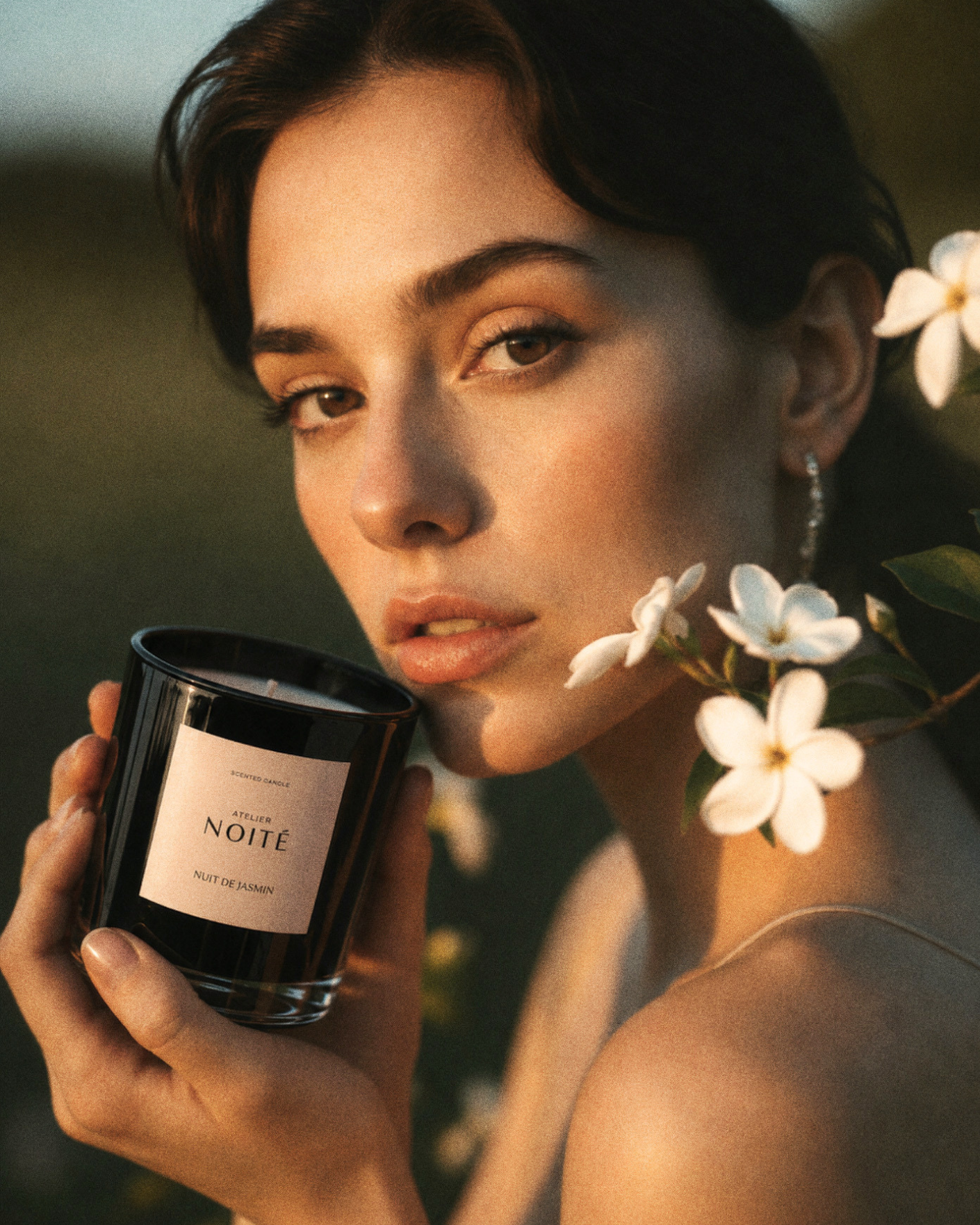 Woman holding a candle labeled 'Atelier Noite' with flowers around her neck.