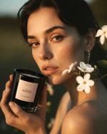 Woman holding a candle labeled 'Atelier Noite' with flowers around her neck.
