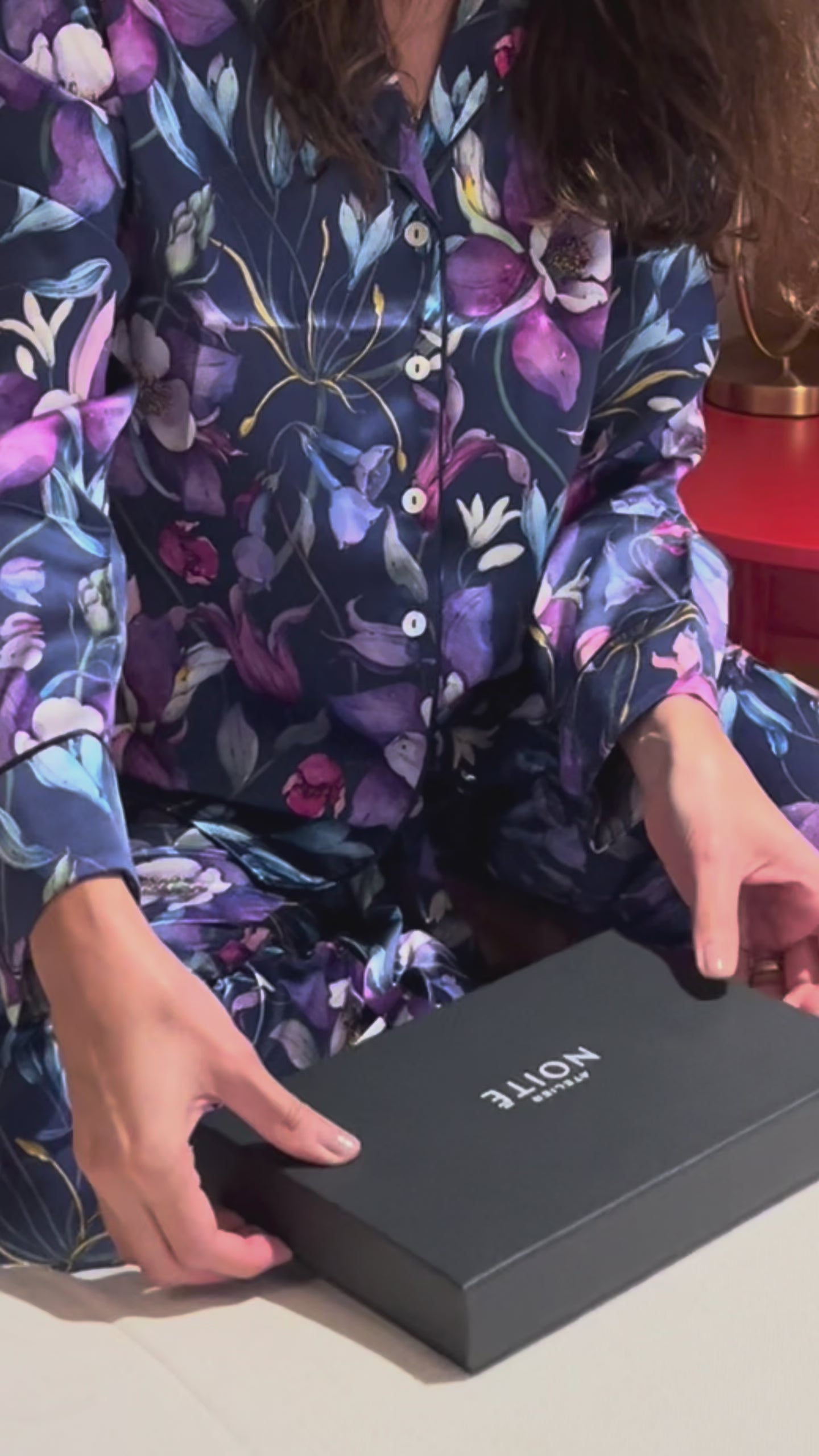 Video of a woman o a bed in a purple pajama opening a gift box containing a purple silk mask