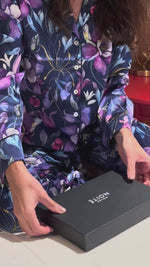 Video of a woman o a bed in a purple pajama opening a gift box containing a purple silk mask